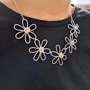 May include: A silver necklace with daisy-shaped pendants. Each daisy has five petals and a coiled centre. The necklace is worn against a black top, which makes the silver metal of the jewellery stand out.