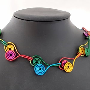 以下が含まれることがあります： ユニークなデザインのカラフルなネックレス。赤、黄、緑、青など、さまざまな色の相互接続された渦巻き状のワイヤーの形が特徴です。 ネックレスは黒いマネキンに展示されています。
