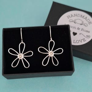 May include: Two silver flower earrings with a wire design, presented in a black box. Each earring features five petals and a coiled centre. A black box with a white label that says "HANDMADE" and "LOVE" is in the background.