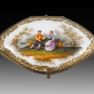May include: A small, oval-shaped porcelain box with a gold-coloured trim and a painted scene of a couple in a landscape. The box has a hinged lid and sits on three small feet.