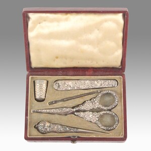 May include: A red velvet box containing a silver sewing kit. The kit includes a thimble, a needle threader, a pair of scissors, a seam ripper, and a small knife.