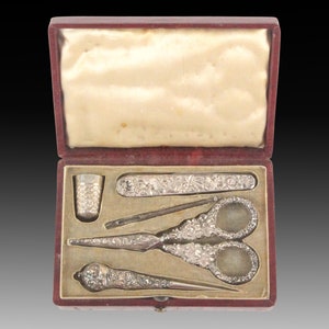 May include: A red velvet box with a white satin lining, containing a silver sewing kit. The kit includes a thimble, a needle threader, a pair of scissors, and a needle case.