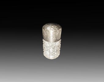 May include: A small, ornate perfume bottle with a silver lid and a clear, textured glass body. The silver lid is decorated with an intricate floral pattern. The glass body has a cut-glass design. The bottle is likely vintage or antique.
