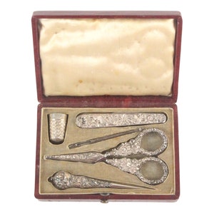 May include: A red velvet box with a white satin lining, containing a silver sewing kit. The kit includes a thimble, a needle threader, a pair of scissors, a seam ripper, and a small knife.