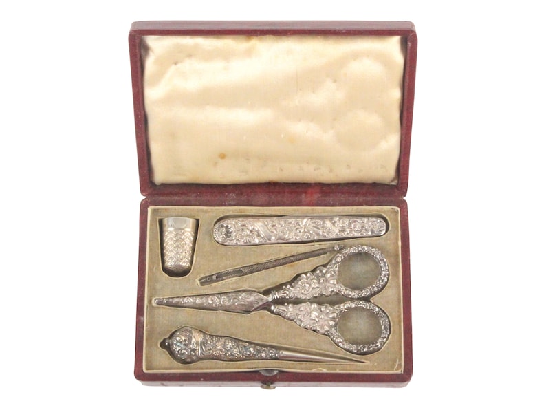 May include: A red velvet box with a white satin lining, containing a silver sewing kit. The kit includes a thimble, a needle threader, a pair of scissors, a seam ripper, and a small knife.