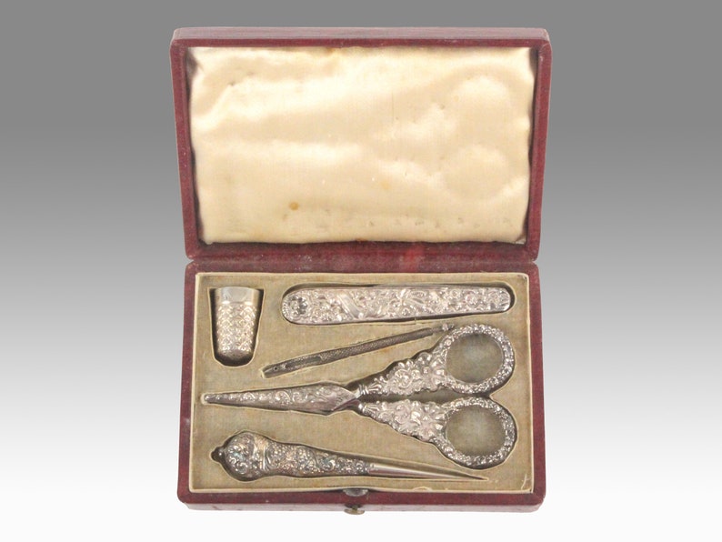 May include: A red velvet box containing a silver sewing kit. The kit includes a thimble, a needle threader, a pair of scissors, a seam ripper, and a small knife.