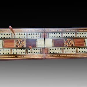 May include: A wooden cribbage board with a marquetry design. The board is open and has two sets of pegs in place. The board is made of different types of wood, including maple, walnut, and cherry.