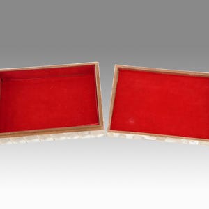 May include: Open rectangular box with a red velvet interior and a mother-of-pearl exterior. The box is divided into two halves, with the lid open, revealing the red lining. The box has a wooden frame.