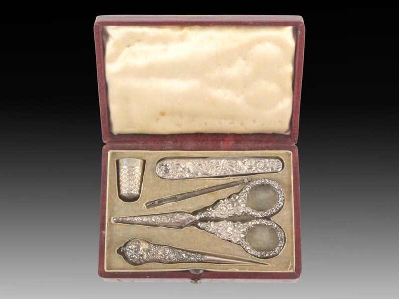 May include: A red velvet box with a white satin lining, containing a silver sewing kit. The kit includes a thimble, a needle threader, a pair of scissors, and a needle case.