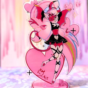 May include: A pink and red acrylic standee featuring a cartoon character with red skin, black clothing, and pink wings. The standee is heart-shaped and has a decorative design. The character has a devilish appearance.