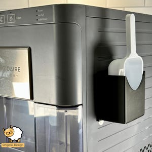 May include: A black and silver refrigerator with a white ice scoop holder attached to the side. The holder has a white scoop inside. The refrigerator has a digital display with the words "Power", "Ice Full", and "Add Water". The refrigerator also has a stainless steel panel with the words "GIGADAIRE GALLERY".