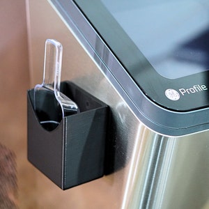 May include: A black plastic scoop holder attached to a stainless steel surface. The holder has a clear plastic scoop inside.