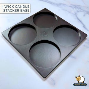 May include: A square black 3 wick candle stacker base with four circular recessed areas for holding candles. The text 3 WICK CANDLE STACKER BASE is visible in the upper left corner, and a small sheep icon with the text Original Design is in the lower right.