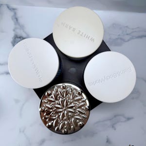 May include: A black plastic candle holder with four circular slots, viewed from above. Three slots feature white lids with the embossed text Bath & Body Works or White Barn. One slot contains a silver metallic lid with a raised floral pattern.