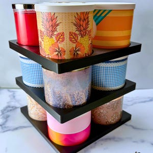 May include: A black tiered candle storage rack consisting of one base and two stackers, designed to hold multiple cylindrical candles. The structure features three levels with circular cutouts for candles, displaying various colourful candle jars.