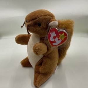 May include: A brown plush squirrel toy with a white belly and a fluffy tail. The toy has a red heart-shaped tag that reads "Beanie Baby". The squirrel is sitting on a white surface.