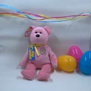 May include: Pink Beanie Baby plush toy with a yellow ribbon and an Easter egg design. The toy is sitting on a white surface with three plastic Easter eggs.