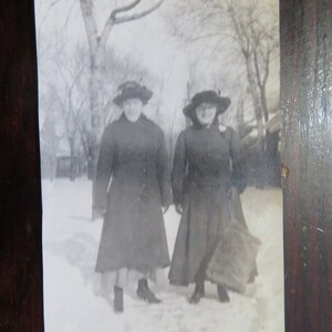 May include: Two women wearing black coats and hats stand in the snow. They are both looking at the camera. The snow is covering the ground and there are trees in the background.