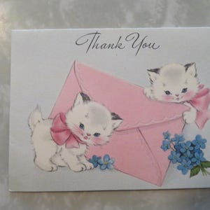 May include: A white rectangular greeting card with the text Thank You in black script. The illustration features two white kittens with pink bows and blue eyes, one standing beside and one peeking over a large pink envelope with blue flowers.