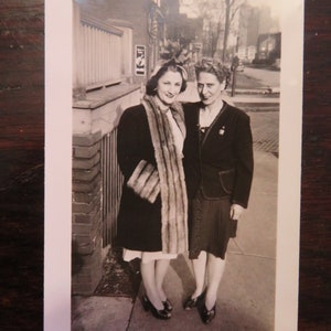 May include: Two women in vintage clothing stand together in front of a brick building. The woman on the left is wearing a black coat with a fur collar and cuffs. The woman on the right is wearing a black dress with a white collar and a black jacket. Both women are smiling.