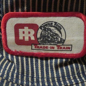 May include: Close-up of a blue and white striped fabric with a red-bordered patch. The patch features a red square with white letters "FR", a black and white train illustration, and the text "POWER TOOL" and "TRADE-IN TRAIN".