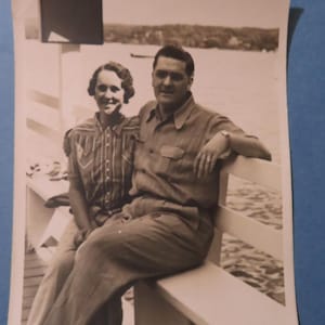 May include: Black and white photograph of a couple sitting on a white bench. The woman wears a patterned shirt and the man wears a button-down shirt. They are posed in front of a body of water with trees in the background.