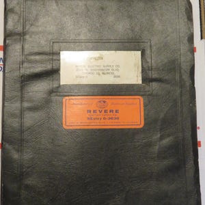 May include: A vintage black binder with two rectangular labels. The top label is white with black text, and the bottom label is orange with white text. The binder is slightly worn.