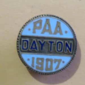 May include: A round, vintage pin with a light blue background and gold lettering. The pin reads "PAA DAYTON 1907" in a circular design. The border is a dark gold color.