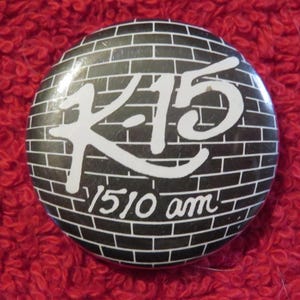 May include: Round button with a black brick wall design. The button features the white text "K-15" and "1510 am" in a stylized font. The button is set against a red, textured background.