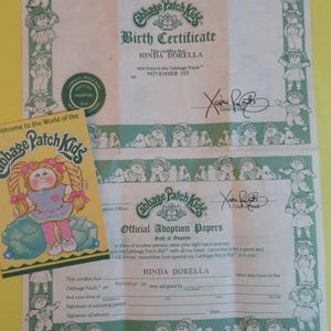 May include: A vintage Cabbage Patch Kids birth certificate and adoption papers, featuring the name Hinda Dorella. The documents are adorned with illustrations of the dolls and the Cabbage Patch Kids logo.