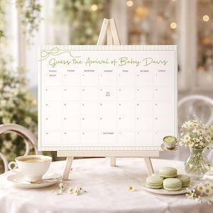 May include: A white calendar on a small wooden easel with the text "Guess the Arrival of Baby Davis" in green script. The calendar grid is empty, with the date "1 September" noted. A teacup, macarons, and flowers are arranged around the easel.