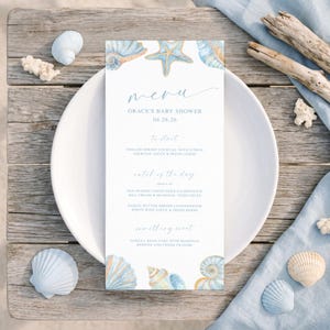 May include: A baby shower menu card with a beach theme. The menu is on a white card with blue watercolor seashells and a starfish design. The menu includes the text "menu", "Grace's Baby Shower", and the date. The card is on a white plate.