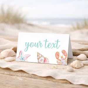 Peut inclure: Carte pliée blanche avec le texte "your text" en écriture turquoise. La carte présente des illustrations à l'aquarelle d'une étoile de mer, d'un coquillage et d'un homard. La carte est sur une surface en bois avec du sable et des coquillages.