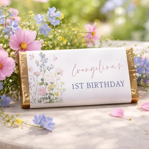 Puede incluir: Una barra de chocolate dorada con una etiqueta blanca que presenta ilustraciones florales y el texto "Evangelina's 1st Birthday". Flores rosas, azules y amarillas rodean la barra, creando un tema festivo.