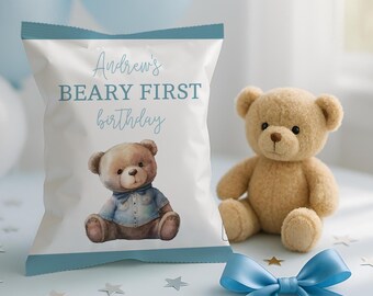 May include: A white snack bag with blue trim featuring the text Andrew's BEARY FIRST birthday and an illustration of a teddy bear wearing a blue shirt. Beside the bag sits a plush tan teddy bear and a blue satin ribbon bow.