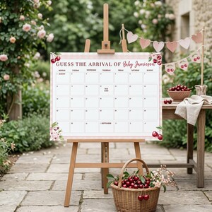 May include: A wooden easel displays a baby arrival calendar with the text "GUESS THE ARRIVAL OF Baby Jamieson." The calendar has a pink and white gingham border and floral accents. A wicker basket filled with cherries sits in front of the easel.