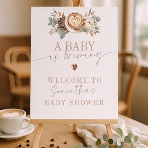 Puede incluir: Un letrero rectangular blanco sobre un caballete de madera con una ilustración en acuarela de una taza de café con arte latte y flores. El texto dice A BABY is brewing, WELCOME TO Samantha's BABY SHOWER, con un icono de grano de café.