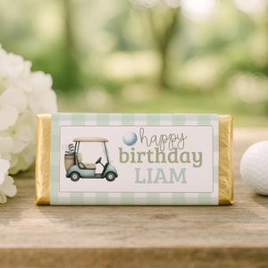 Pode incluir: Uma barra de chocolate retangular embrulhada em papel dourado com um rótulo xadrez verde claro e branco. O rótulo apresenta uma ilustração de um carrinho de golfe, uma bola de golfe e o texto "happy birthday LIAM" em fontes variadas.