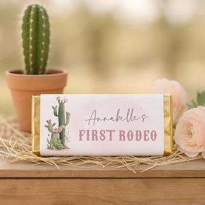 May include: A rectangular chocolate bar wrapped in white paper with gold foil ends. The label features a watercolour illustration of a green cactus with pink flowers and the text "Annabelle's FIRST RODEO" in black and dusty pink lettering.