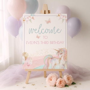 Op de afbeelding: Een rechthoekig verjaardagsbord op een houten ezel met een eenhoornillustratie, pastelkleurige ballonnen, vlinders en vlaggetjes. De tekst luidt: welcome TO EVELYN'S THIRD BIRTHDAY. Het ontwerp bevat gouden glitteraccenten en zachte roze tinten.