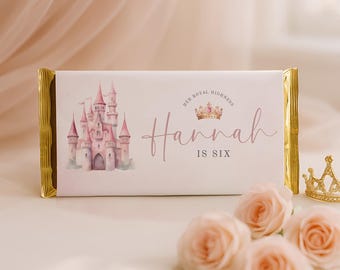 Könnte beinhalten: Eine rechteckige Schokoladenverpackung mit einer Aquarellillustration eines rosa Schlosses auf der linken Seite und dem Text "Her Royal Highness Hannah is six" mit einer kleinen Kronengrafik in der Mitte, abgerundet mit goldener Folie an den Enden.