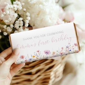 Op de afbeelding: Een rechthoekige chocoladewikkel met een witte achtergrond en onderaan een delicate aquarelillustratie van wilde bloemen. De tekst luidt THANK YOU FOR CELEBRATING Emma's first birthday in donkergrijze en lichtroze letters.