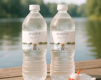 May include: Two clear plastic water bottles with white caps, each featuring a custom label that reads DAISY'S baby shower above an illustration of a person in a straw hat fishing on a dock. The labels have a soft, watercolor-style lake scene.