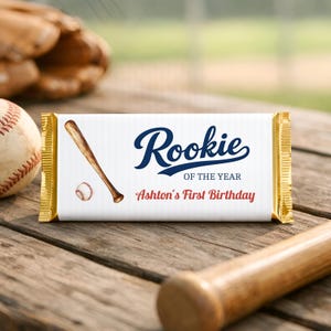 May include: A rectangular candy bar wrapper with gold foil ends, featuring a white background with a baseball bat and ball illustration. The text reads Rookie OF THE YEAR and Ashton's First Birthday in navy blue and red lettering.