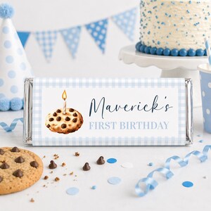 Puede incluir: Envoltorio de barra de caramelo a cuadros azules y blancos con el texto "Maverick's FIRST BIRTHDAY" y una ilustración de galleta con una vela encendida. Una tarta de cumpleaños, un gorro de fiesta y confeti están en el fondo.