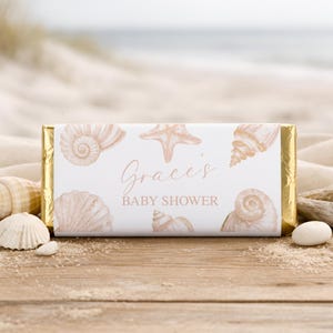 May include: A white candy bar wrapper with gold foil edges, decorated with seashell illustrations and the text "Grace's BABY SHOWER". The design features starfish and spiral shells in a soft, neutral colour palette.