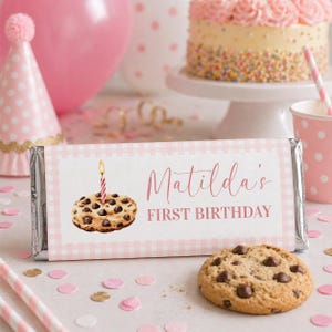 May include: A candy bar wrapper with a pink gingham pattern, featuring the text "Matilda's FIRST BIRTHDAY" and an illustration of a chocolate chip cookie with a lit candle. Pink balloons and a cake are in the background.