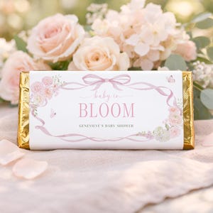 May include: A chocolate bar wrapped in gold foil with a white label. The label features the text "baby in BLOOM" in pink script, surrounded by floral and ribbon designs. The text "GENEVIEVE'S BABY SHOWER" is also present.