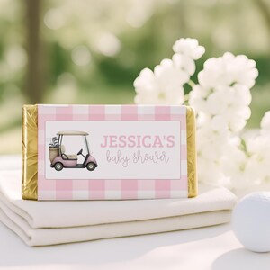 Pode incluir: Uma barra retangular embrulhada em papel alumínio dourado com um rótulo xadrez rosa e branco. O rótulo apresenta uma ilustração de um carrinho de golfe rosa e o texto JESSICA'S baby shower em letras rosa e cinza.