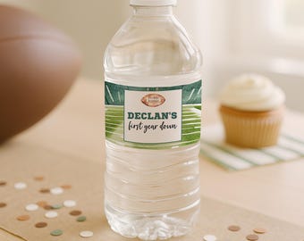 May include: A clear plastic water bottle with a custom label featuring a green football field graphic, a small brown football icon, and the text "DECLAN'S first year down" printed in dark green and black lettering on a white background.